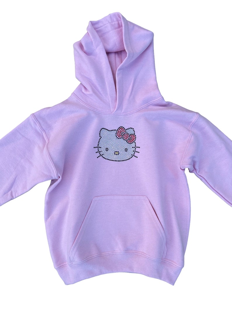 Puede incluir: Una sudadera con capucha rosa claro con un dise&ntilde;o de Hello Kitty de diamantes de imitaci&oacute;n. La sudadera tiene un bolsillo canguro y una capucha con cord&oacute;n.