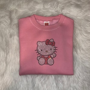 K&ouml;nnte beinhalten: Rosafarbener Pullover mit einem Strass-Hello-Kitty-Motiv.