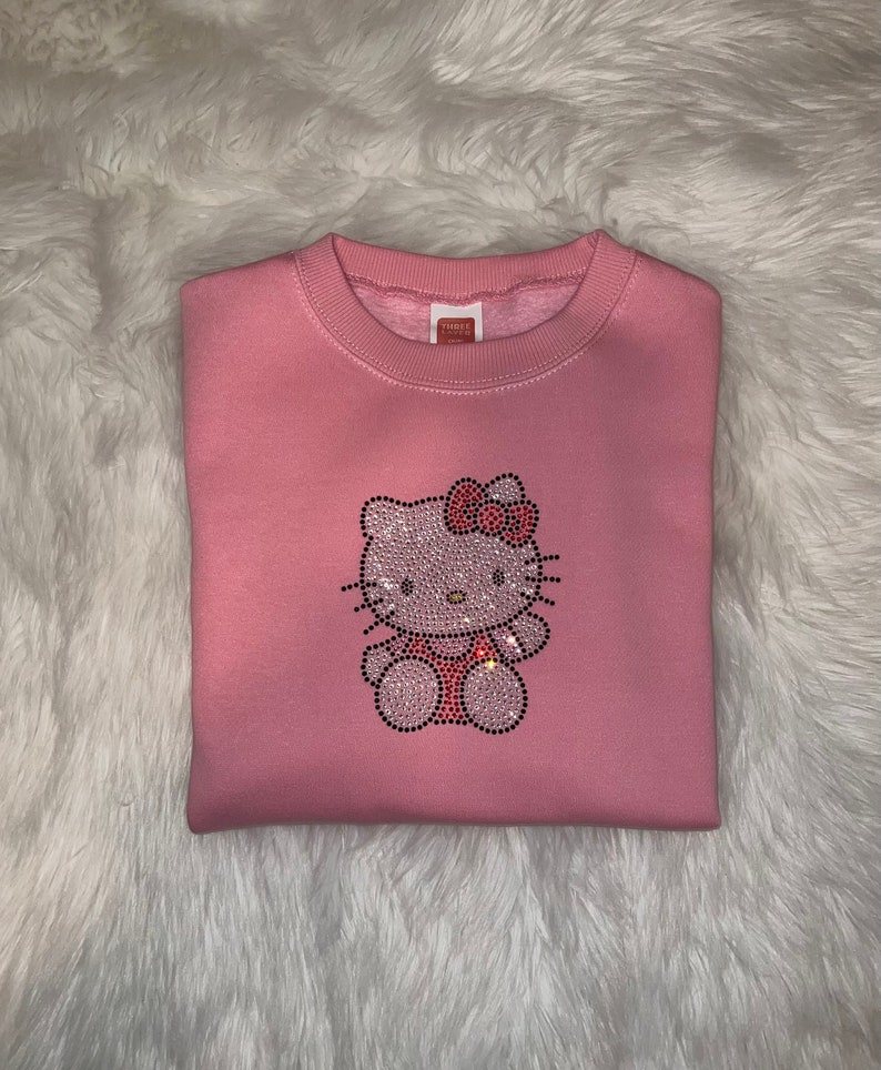 K&ouml;nnte beinhalten: Rosafarbener Pullover mit einem Strass-Hello-Kitty-Motiv.