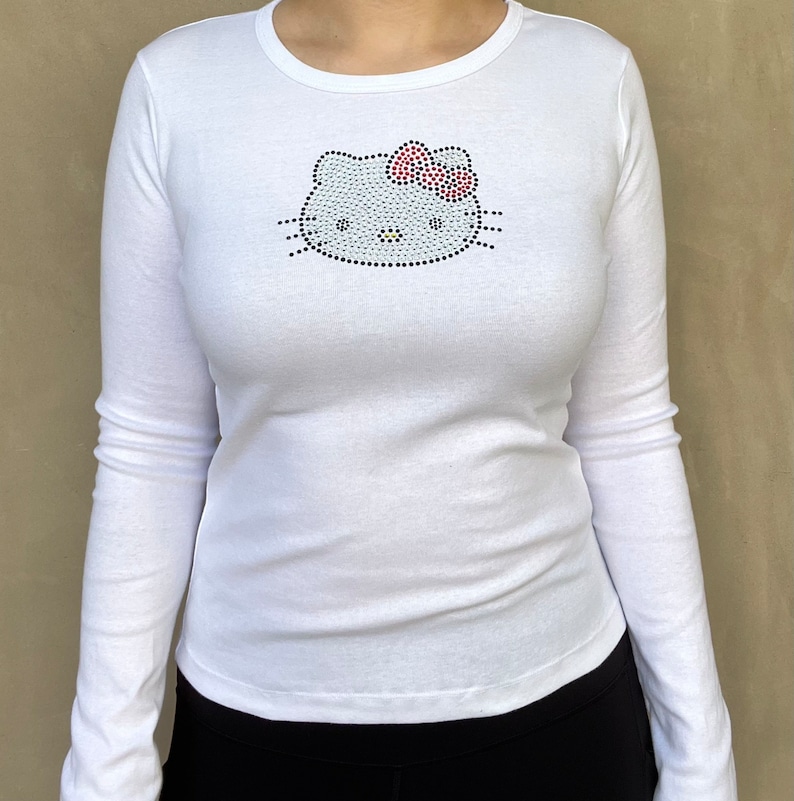 May include: White long-sleeved shirt with a rhinestone Hello Kitty design on the front.