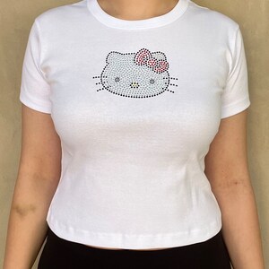 Pu&ograve; includere: Maglietta bianca con un design Hello Kitty in strass.