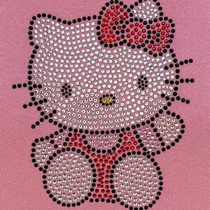 K&ouml;nnte beinhalten: Ein rosa Stoff mit einem Strass-Hello-Kitty-Design. Das Design zeigt eine wei&szlig;e Hello Kitty mit einer roten Schleife und einem schwarzen Umriss.