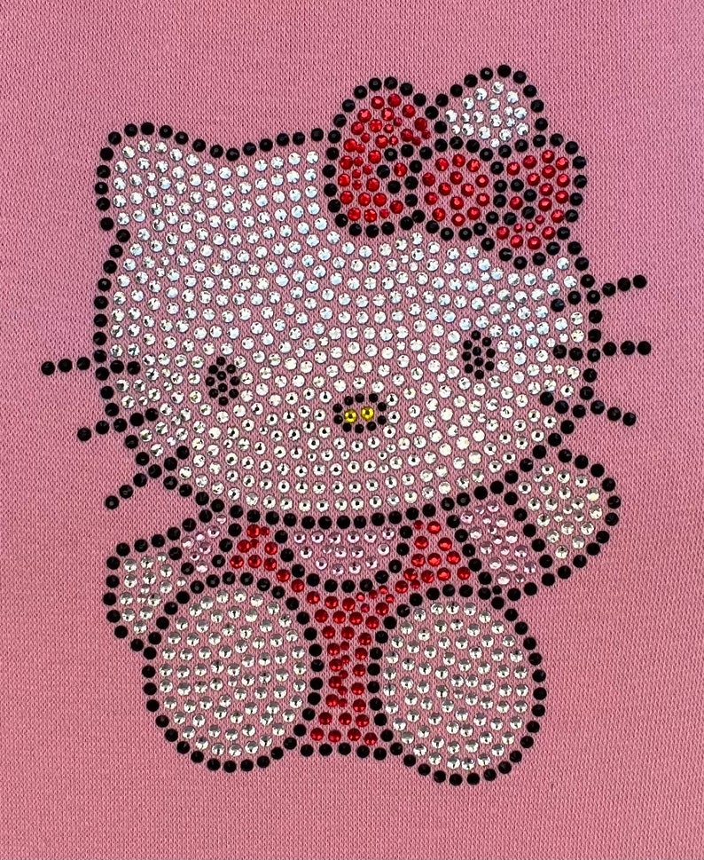 K&ouml;nnte beinhalten: Ein rosa Stoff mit einem Strass-Hello-Kitty-Design. Das Design zeigt eine wei&szlig;e Hello Kitty mit einer roten Schleife und einem schwarzen Umriss.