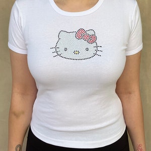 Puede incluir: Camiseta blanca con un dise&ntilde;o de Hello Kitty de pedrer&iacute;a. El dise&ntilde;o presenta una cara de Hello Kitty blanca con un lazo rojo.