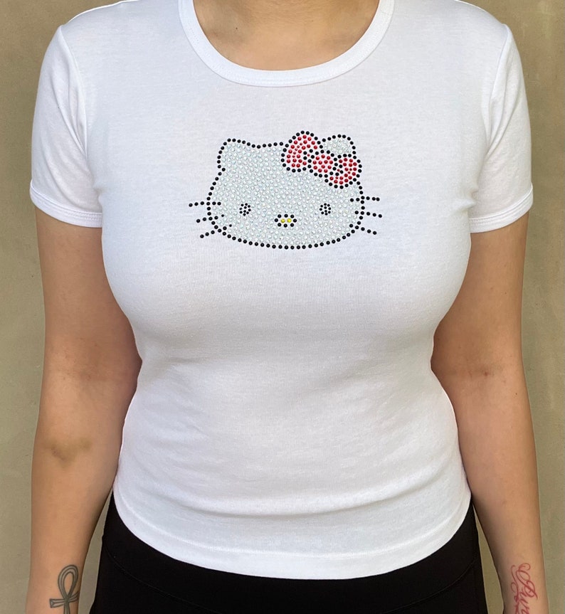 Puede incluir: Camiseta blanca con un dise&ntilde;o de Hello Kitty de pedrer&iacute;a. El dise&ntilde;o presenta una cara de Hello Kitty blanca con un lazo rojo.