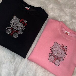 K&ouml;nnte beinhalten: Zwei Sweatshirts mit einem Strass-Hello-Kitty-Design. Ein Sweatshirt ist schwarz und das andere ist rosa.