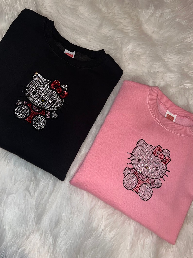 K&ouml;nnte beinhalten: Zwei Sweatshirts mit einem Strass-Hello-Kitty-Design. Ein Sweatshirt ist schwarz und das andere ist rosa.