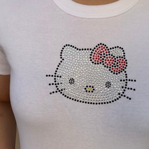 Pu&ograve; includere: Maglietta bianca con un design Hello Kitty in strass. Il fiocco &egrave; rosso e il resto del design &egrave; in strass trasparente.