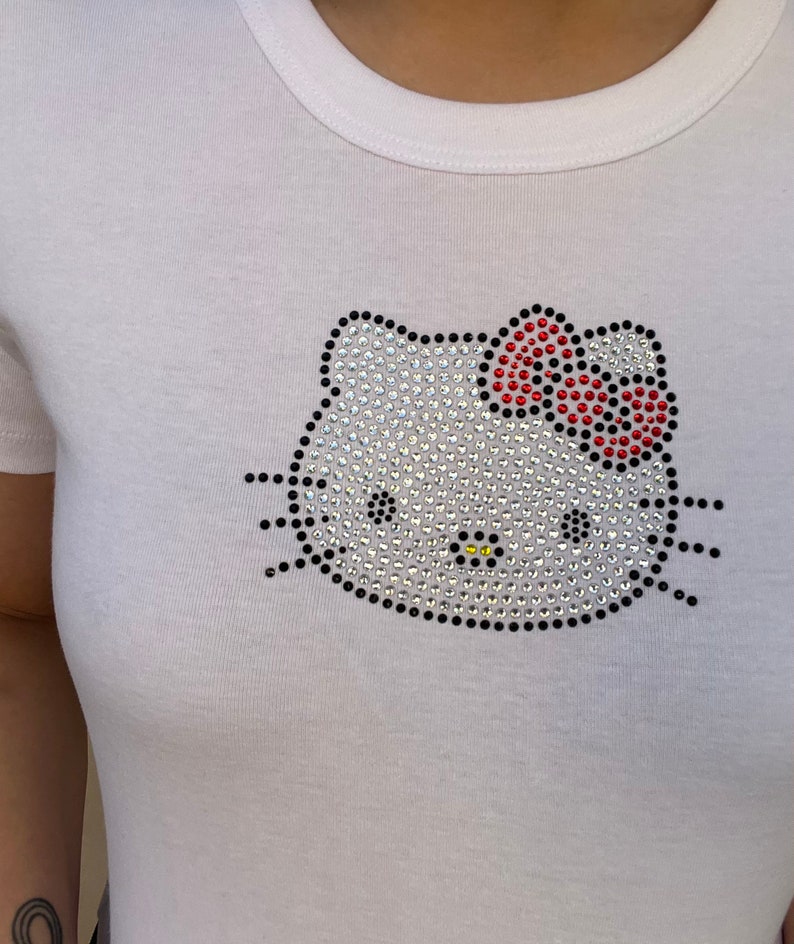 Pu&ograve; includere: Maglietta bianca con un design Hello Kitty in strass. Il fiocco &egrave; rosso e il resto del design &egrave; in strass trasparente.