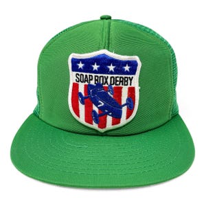 May include: Green trucker hat with a curved brim and mesh back. The front features a patch with the text "SOAP BOX DERBY" and a blue race car design on a red, white, and blue shield.