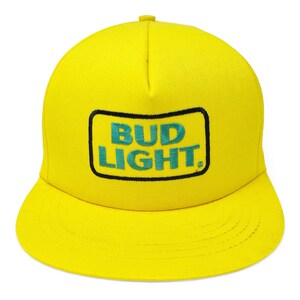 May include: A bright yellow, flat-brimmed baseball cap. The cap features the text "BUD LIGHT" in teal lettering within a black-outlined rectangle on the front. The cap is a solid yellow color.