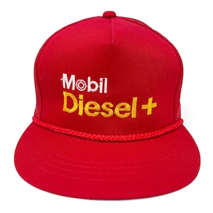 Könnte beinhalten: Rote Trucker-Cap mit dem Schriftzug "Mobil Diesel +" in Weiß und Gelb. Die Kappe hat einen roten Schirm und eine rote Mesh-Rückseite. Ein rotes Seildetail verläuft entlang des Schirms.