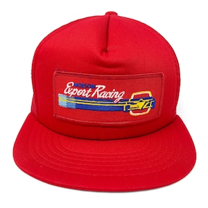 May include: A red trucker hat with a mesh back and a curved brim. The front features a rectangular patch with the text "Expert Racing" in white script, along with a blue, yellow, and white design.