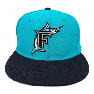 May include: A teal and navy blue baseball cap featuring the letter "F" with a marlin fish logo. The cap has a flat brim and a structured crown. The cap is a sports-themed accessory.