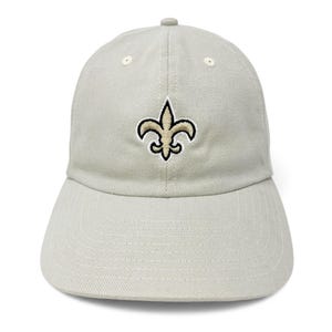 May include: A light gray baseball cap featuring the New Orleans Saints fleur-de-lis logo embroidered on the front. The logo is in gold with a black outline. The cap has a curved brim and an adjustable strap.