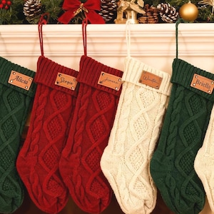 Könnte beinhalten: Sechs Zopfstrick-Weihnachtsstrümpfe in Creme, Grün und Rot, jeweils mit einem Namensschild aus Holz, hängen über einem Kamin. Der Kamin hat ein glühendes Feuer und ist mit festlichem Grün und Ornamenten geschmückt.