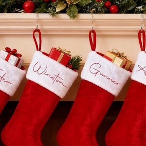 Könnte beinhalten: Vier rote Weihnachtsstrümpfe mit weißen Bündchen und den Namen Tadeo, Winston, Gunner und Armani in Schreibschrift, hängen über einem Kamin. Jeder Strumpf ist mit Geschenken und Grünzeug gefüllt.