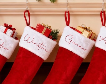 May include: Four red Christmas stockings hang from a fireplace mantel, each with a white cuff and a name embroidered in cursive: Tadeo, Winston, Gunner, and Armani. Each stocking is filled with gifts and greenery, creating a festive holiday scene.