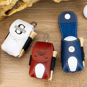 Könnte beinhalten: Drei personalisierte Golfballhalter aus weißem, rotem und blauem Leder. Jeder Halter enthält einen Golfball und Tees. Der weiße Halter ist mit "STEPHEN" beschriftet, der rote Halter mit "CARLOS". Ein Golfball und ein Pitchgabel sind ebenfalls sichtbar.