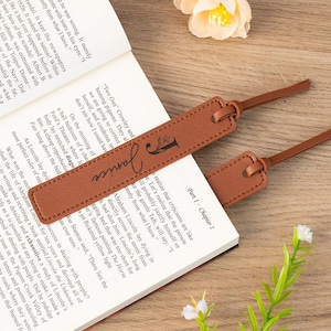 Könnte beinhalten: Zwei braune Leder-Lesezeichen mit dem Namen "Janice" und dem Buchstaben "J" aufgedruckt, die auf einem offenen Buch liegen. Die Lesezeichen haben eine genähte Bordüre und einen dünnen Riemen.