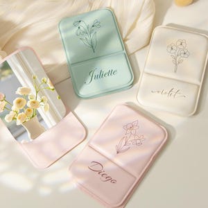 Könnte beinhalten: Vier kleine, rechteckige Kompaktspiegel in Pastellfarben. Jeder Spiegel hat ein Blumendesign und einen Namen: Juliette, Violet und Diego. Ein Spiegel reflektiert eine Blumenvase. Make-up-Pinsel sind im Hintergrund.
