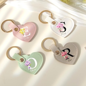 Può includere: Quattro portachiavi a forma di cuore in rosa, bianco, verde e grigio. Ogni portachiavi presenta un anello dorato, un cinturino in pelle e un design floreale con una lettera e un nome. Le lettere sono E, S, O e R.