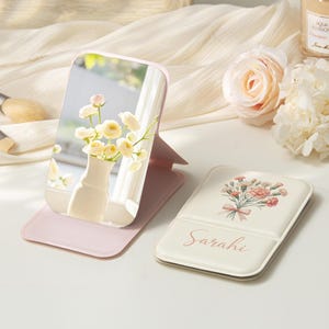 Könnte beinhalten: Ein pinkfarbener, faltbarer Schminkspiegel mit rechteckiger Spiegelfläche. Der Spiegel reflektiert eine Vase mit gelben Blumen. Ein passendes Etui mit Blumendesign und dem Namen "Sarah" ist ebenfalls vorhanden.