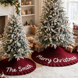 May include: Two round, deep red knitted Christmas tree skirts with ruffled edges. One skirt features the white embroidered text 'The Smith' and the other features the white embroidered text 'Merry Christmas' placed around the base of two flocked trees.