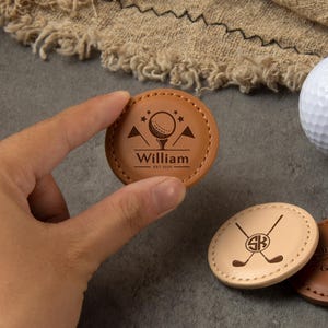 Könnte beinhalten: Eine Sammlung personalisierter Golfballmarker aus Leder in verschiedenen Braun- und Schwarztönen, mit einem weißen Golfball. Ein Marker wird von einer Hand gehalten. Die Marker weisen eingravierte Designs auf, darunter ein Golfball, gekreuzte Schläger und Text.
