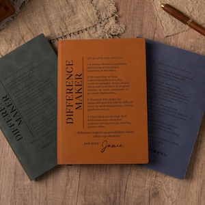 Puede incluir: Tres cuadernos con las palabras "Difference Maker" en la portada. Los cuadernos son en tonos de verde, naranja y azul. El cuaderno naranja tiene texto en la portada y el nombre "Jamie" en la parte inferior. También se ven un bolígrafo y una nota de "Thank You".