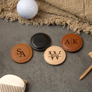 Puede incluir: Una composición temática de golf con una pelota blanca, marcadores de bola de cuero personalizados y tees de madera. Los marcadores son de varios colores, grabados con iniciales y nombres. También se ve un palo de golf.