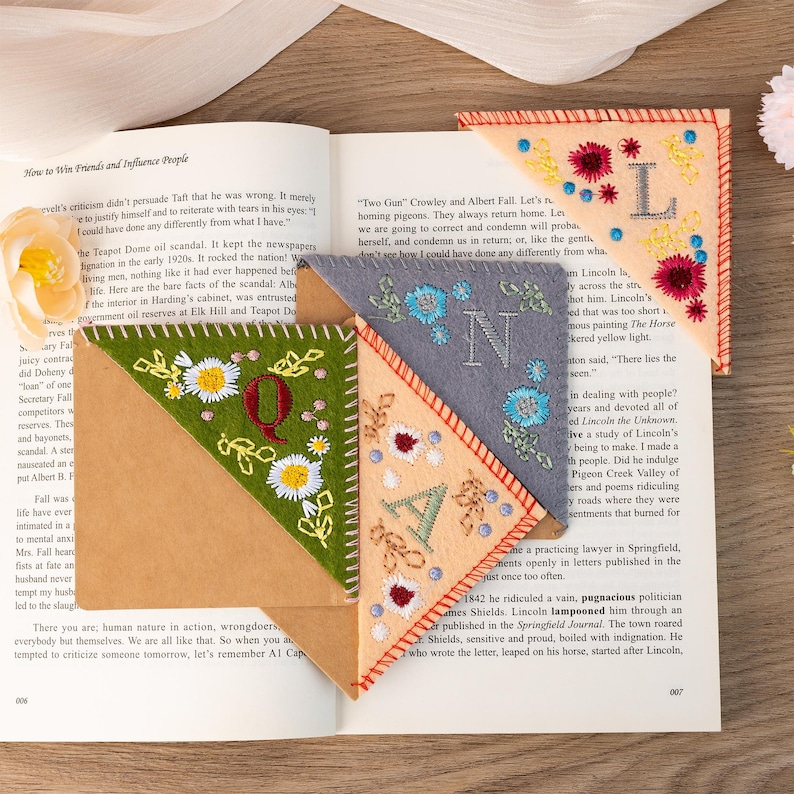 May include: Four felt corner bookmarks with embroidered letters, flowers, and stars. The letters are Q, N, A, and L. The bookmarks are in shades of green, brown, gray, and peach.