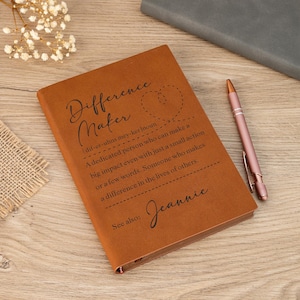 Puede incluir: Un diario de cuero marrón con las palabras "Difference Maker" y una definición impresa en la portada. Un bolígrafo de oro rosa descansa junto al diario sobre una superficie de madera. El diario incluye el nombre "Jeannie".
