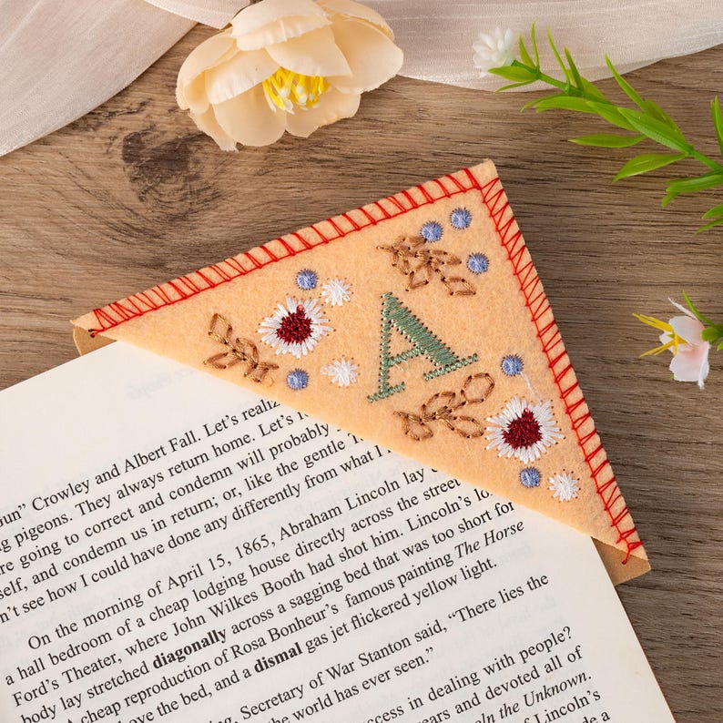 May include: A peach-colored felt bookmark with a large embroidered letter "A" in green and white. The bookmark is decorated with small embroidered flowers in red, white, and blue. The bookmark is folded over a book.