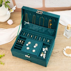 May include: A green velvet jewelry box with a hinged lid and a silver lock. The box has multiple compartments for storing rings, earrings, and necklaces. The box is open and the compartments are filled with jewelry.