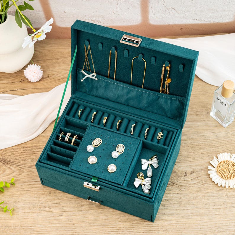 May include: A green velvet jewelry box with a hinged lid and a silver lock. The box has multiple compartments for storing rings, earrings, and necklaces. The box is open and the compartments are filled with jewelry.