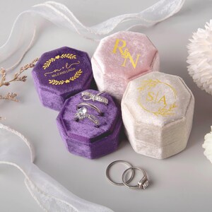 May include: An assortment of velvet ring boxes in shades of purple, pink, and cream. The boxes are hexagonal and feature gold lettering and designs. Two rings are displayed in one of the purple boxes. The text on the boxes reads "Milo and Linda", "R&N", and "S&A".