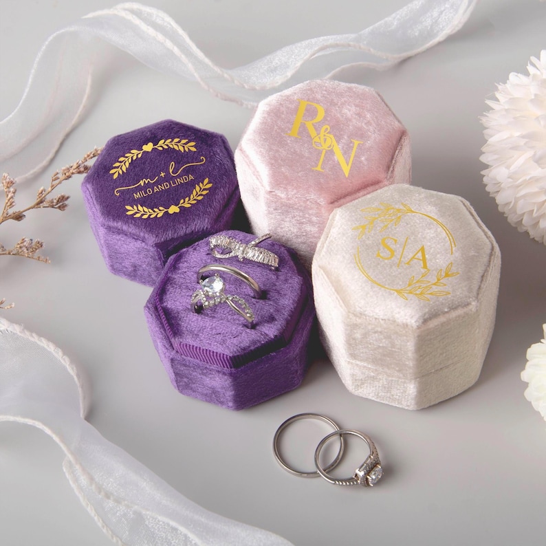May include: An assortment of velvet ring boxes in shades of purple, pink, and cream. The boxes are hexagonal and feature gold lettering and designs. Two rings are displayed in one of the purple boxes. The text on the boxes reads "Milo and Linda", "R&N", and "S&A".