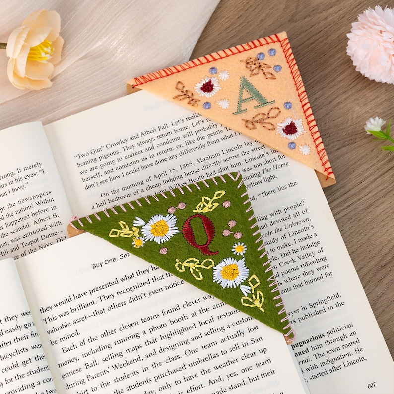 May include: Two felt corner bookmarks with embroidered letters and floral designs. The first bookmark is peach with the letter "A" and the second bookmark is green with the letter "Q". Both bookmarks have a white stitched border.