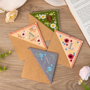 May include: Four handmade felt bookmarks with embroidered letters and floral designs. The bookmarks are in the shape of a triangle and are made of brown felt with a contrasting felt triangle on top. The letters are A, L, N, and Q. The bookmarks are on a wooden surface.