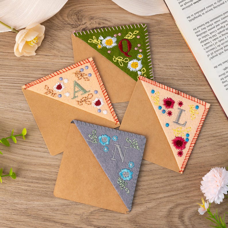 May include: Four handmade felt bookmarks with embroidered letters and floral designs. The bookmarks are in the shape of a triangle and are made of brown felt with a contrasting felt triangle on top. The letters are A, L, N, and Q. The bookmarks are on a wooden surface.