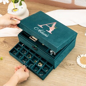 May include: A three-tiered jewelry box with a teal velvet exterior. The top tier has a hinged lid with a personalized inscription that reads "Alexis" and a pink floral letter "A". The bottom two tiers have pull-out drawers with compartments for rings, earrings, and other jewelry.