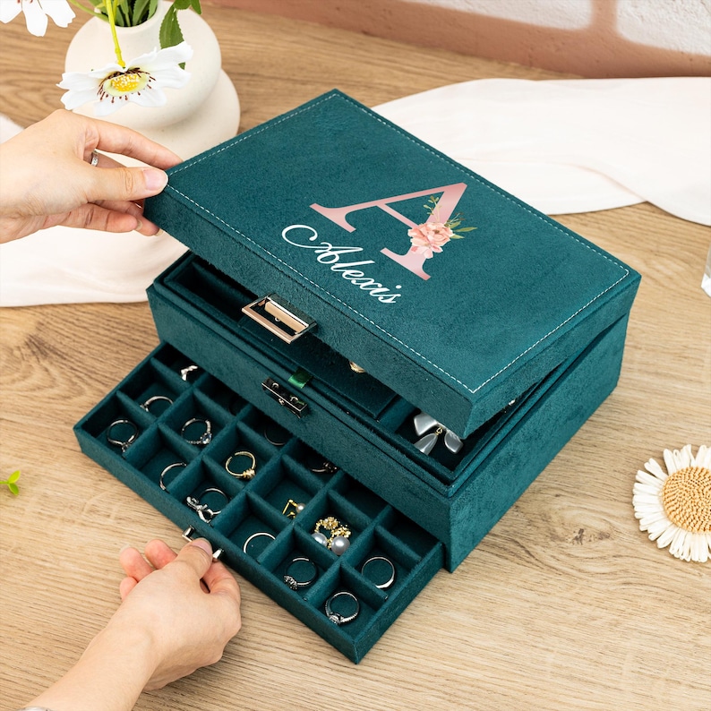 May include: A three-tiered jewelry box with a teal velvet exterior. The top tier has a hinged lid with a personalized inscription that reads "Alexis" and a pink floral letter "A". The bottom two tiers have pull-out drawers with compartments for rings, earrings, and other jewelry.