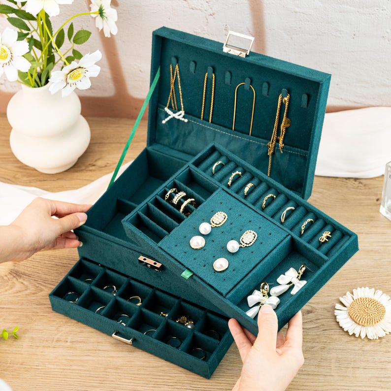 May include: A three-tiered green velvet jewelry box with a lock and key. The top tier has a hinged lid with slots for necklaces and earrings. The middle tier has compartments for rings and earrings. The bottom tier has a drawer with compartments for rings and other small jewelry.