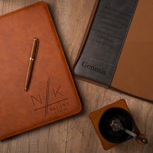 Könnte beinhalten: Ein braunes Lederjournal mit den Initialen "N/K" und dem Namen "Kiara Nico" wird neben einem Stift gezeigt. Ein weiteres Journal mit dem Namen "Genesis" ist sichtbar. Eine hölzerne Kaffeemühle ist ebenfalls vorhanden. Das Bild enthält Text: "Eine engagierte Person, die einen großen Einfluss haben kann..."