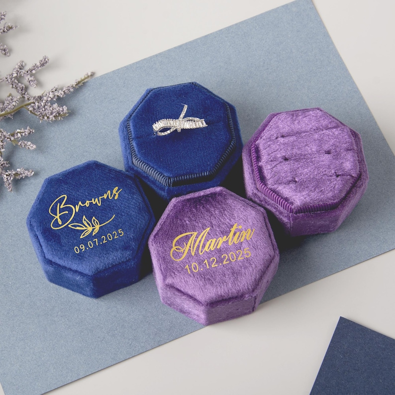 May include: Four velvet ring boxes in shades of blue and purple. One blue box holds a ring, while the others are empty. Two boxes have gold text with names and dates. The boxes are octagonal and sit on a blue surface.