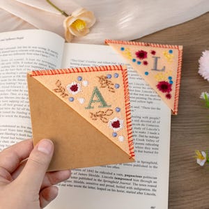 May include: Two felt corner bookmarks with embroidered letters A and L. The bookmarks are peach colored with white and red floral designs. The bookmarks are on top of a brown book.
