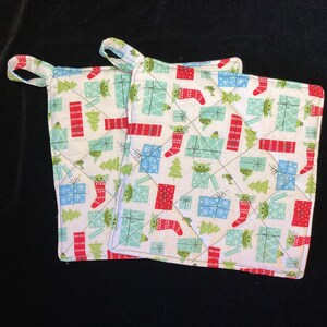 May include: Two white quilted pot holders with a Christmas theme. The fabric features a repeating pattern of green trees, red and blue wrapped presents, and a green alien character wearing a red stocking cap.