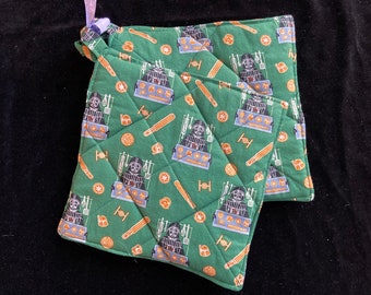Könnte beinhalten: Zwei grüne, gesteppte Topflappen mit einem dunkelgrünen und schwarzen Muster von Darth Vader und Star Wars-Motiven. Die Topflappen haben eine Schlaufe zum Aufhängen.