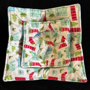 May include: Set of two quilted bowl cozies. The cozies feature a white background with a repeating pattern of green Christmas trees, red wrapped gifts, and a green character. The edges are trimmed with a light blue binding.